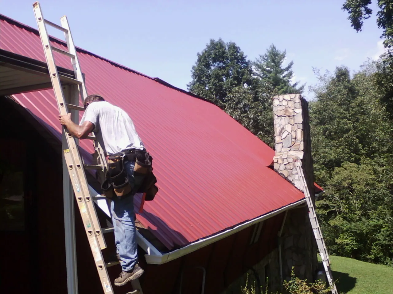 Roofer on a ladder beside a red metal roof and stone chimney — Soffit & Fascia Repair by Roofing Brothers in East Bernard
